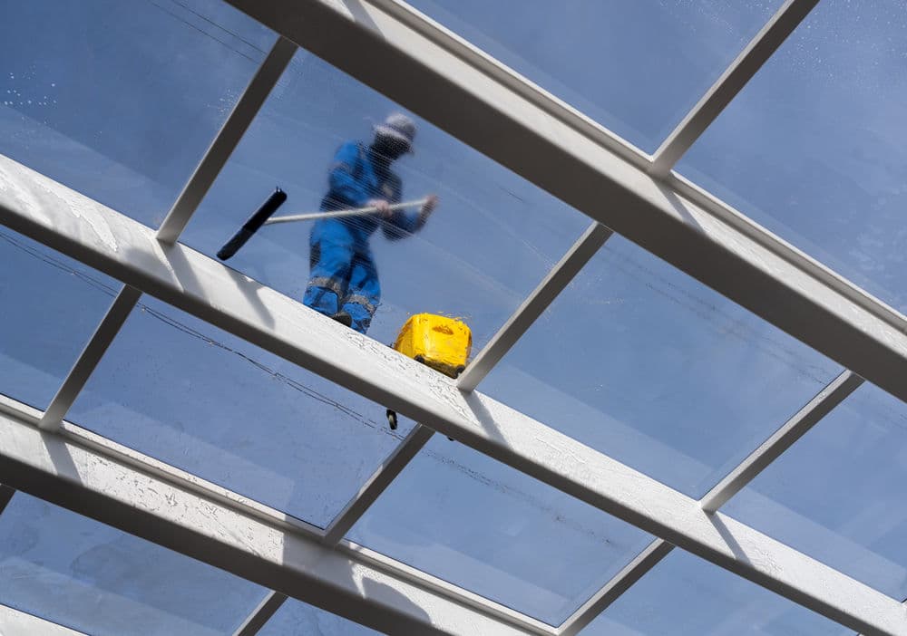 Person cleaning a glass roof with a squeegee and yellow bucket against a blue sky.